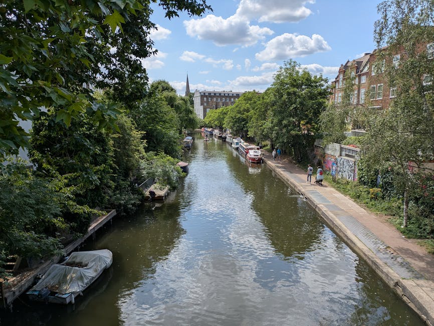 A brown wooden houseboat named Poppy is moored on a canal with calm water reflecting its image and the surrounding buildings. The boat features a covered seating area with a red roof, metal railings, 